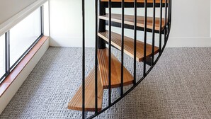 Una escalera de caracol con escalones de madera y barandillas de metal negro en una habitación con ventanas amplias y piso alfombrado.