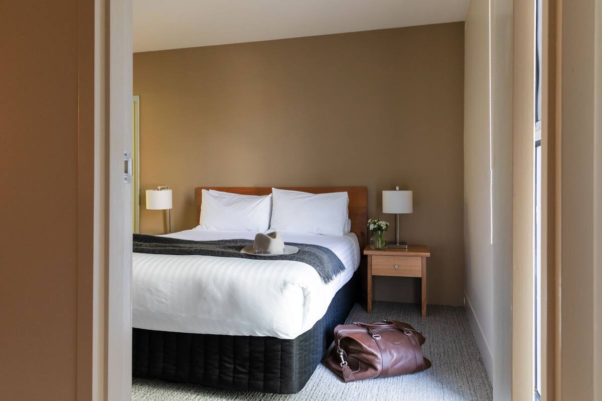 A hotel room with a bed, bedside lamps, a nightstand, and a handbag on the floor.