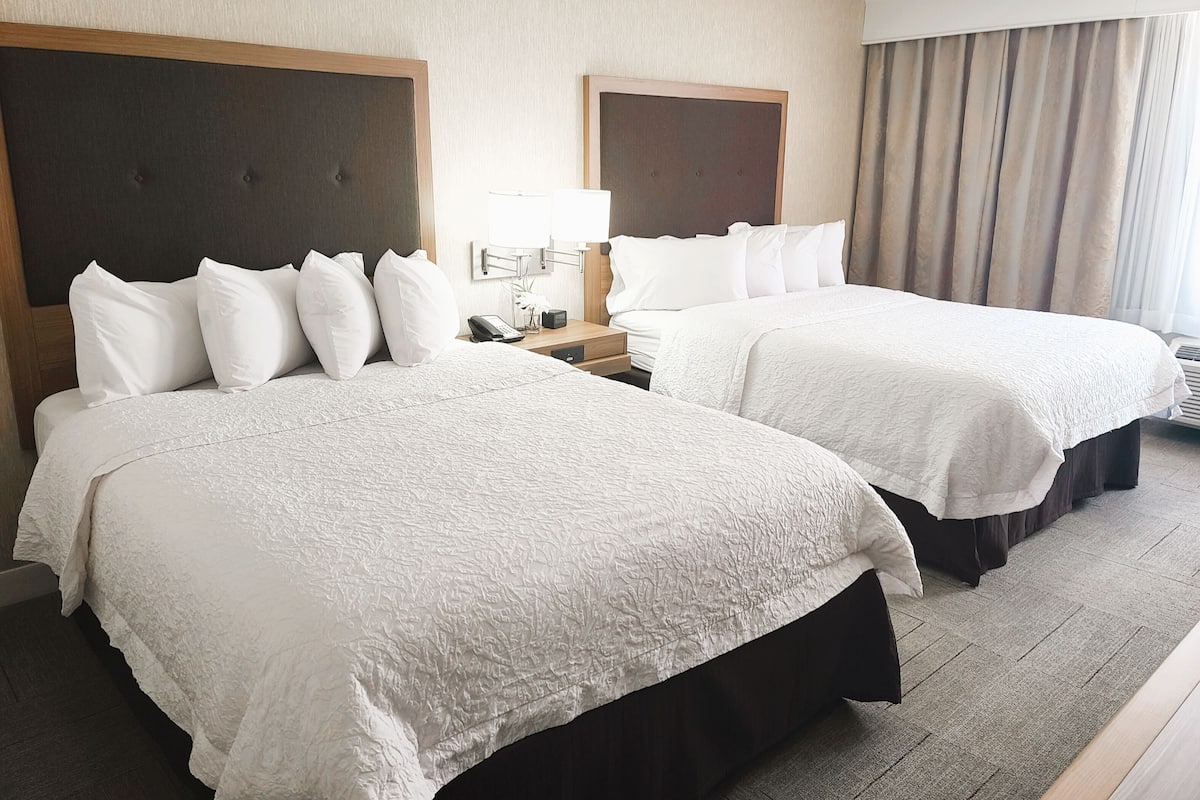 A hotel room with two beds, each with white linens and a headboard. There is a bedside table with a lamp and a phone. The room has a neutral color scheme with beige walls and curtains.