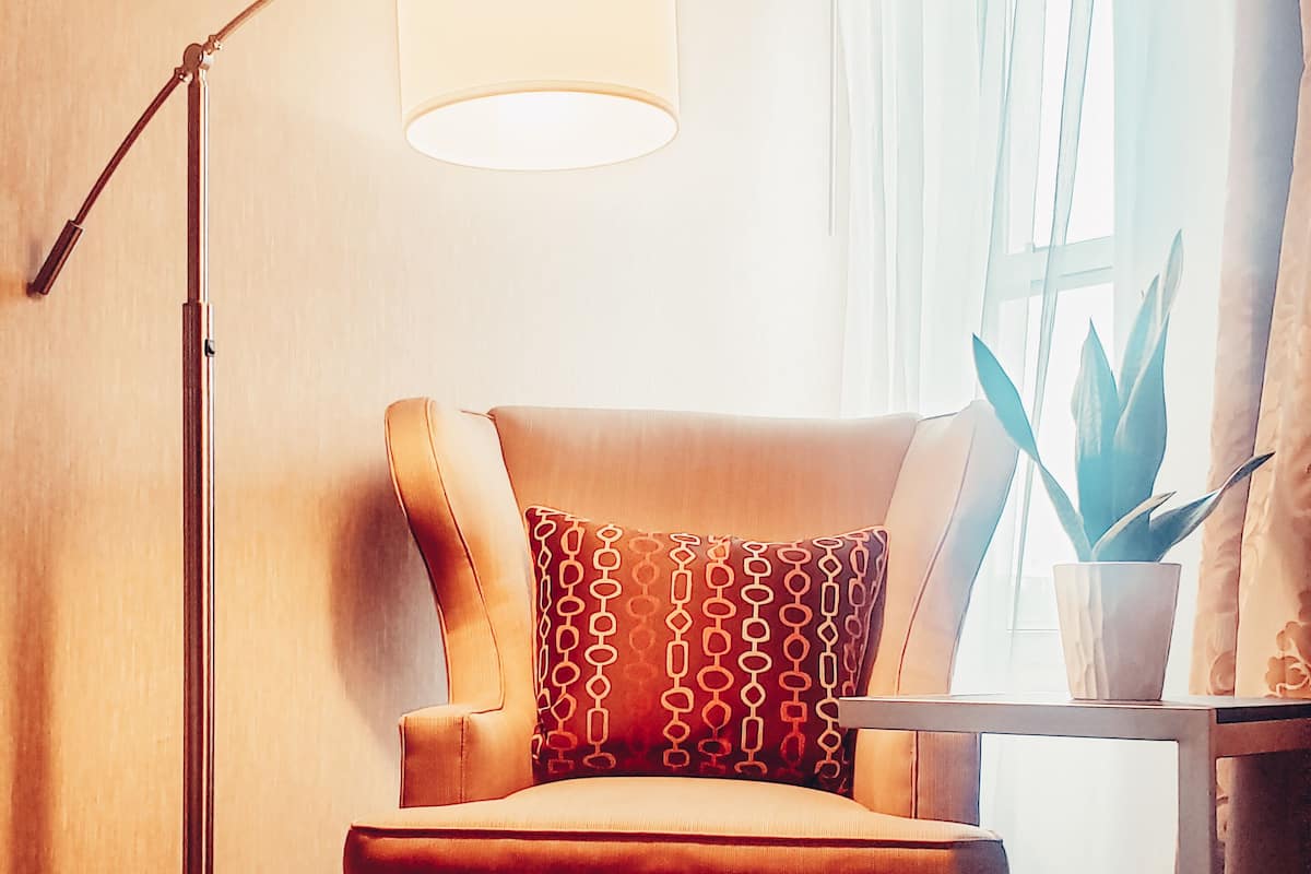 A corner room with a brown leather armchair, a floor lamp, a small table with a potted plant, and a window with sheer curtains.