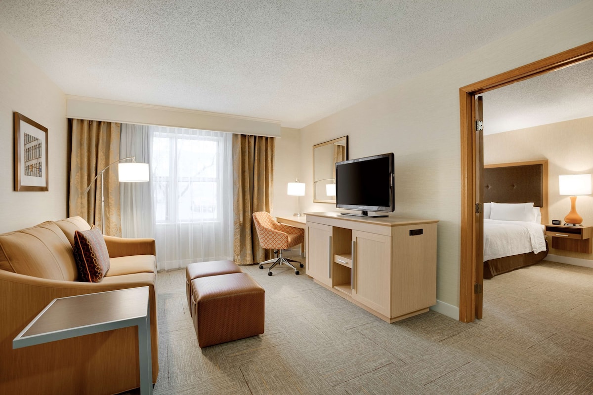 A hotel room with a sofa, a desk with a TV, a chair, a bed, and a bedside table.