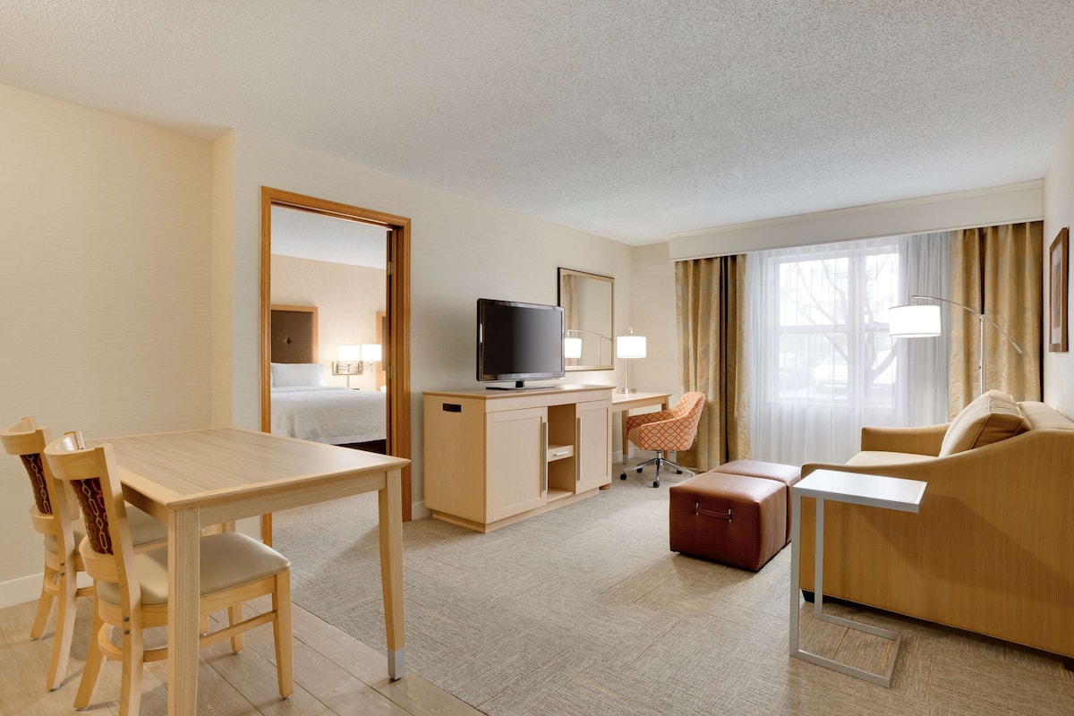 A hotel room with a wooden dining table, a TV on a cabinet, a sofa, a desk, and a chair.