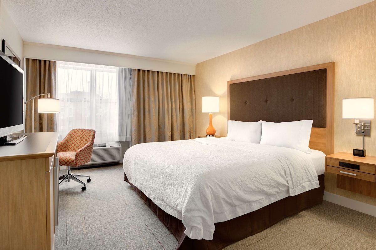 A hotel room with a large bed, a desk, a chair, and a window with curtains.