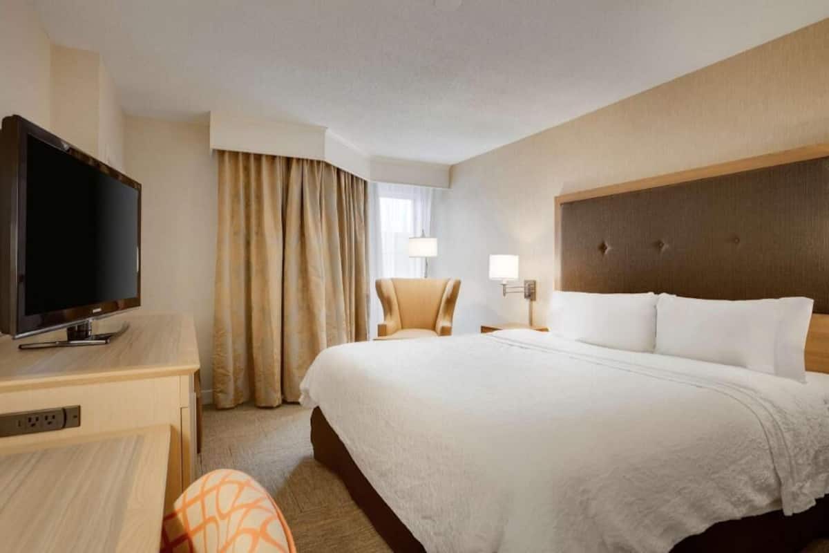 A hotel room with a bed, a desk with a television, a chair, a lamp, and a window with curtains.