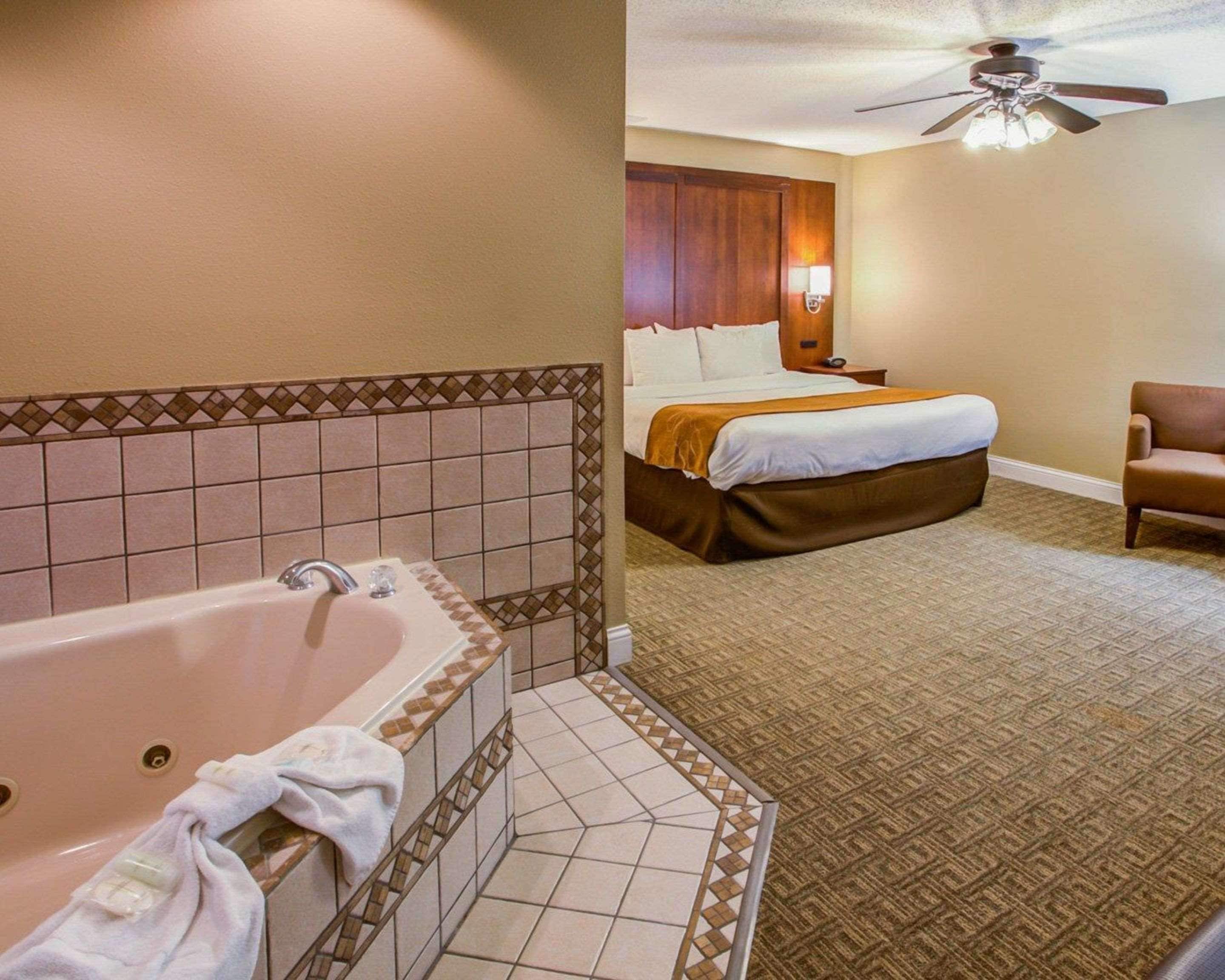 A hotel room with a large bathtub, a bed with a wooden headboard, a ceiling fan, and a patterned carpet.