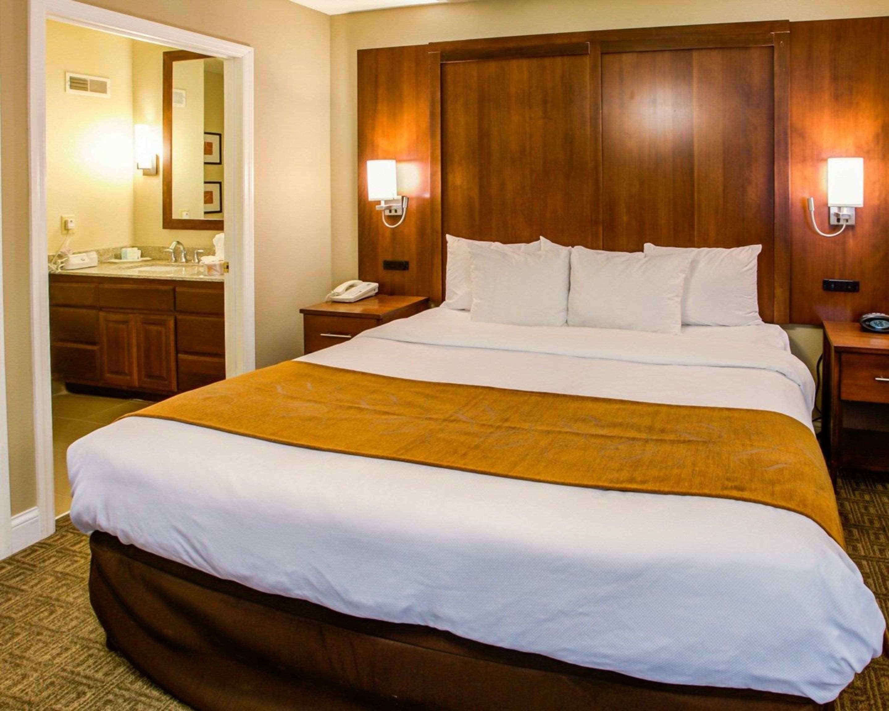Een hotelkamer met een groot bed, een houten hoofdeinde, twee bedlampen, een nachtkastje, een telefoon en een badkamer die door een open deur zichtbaar is.