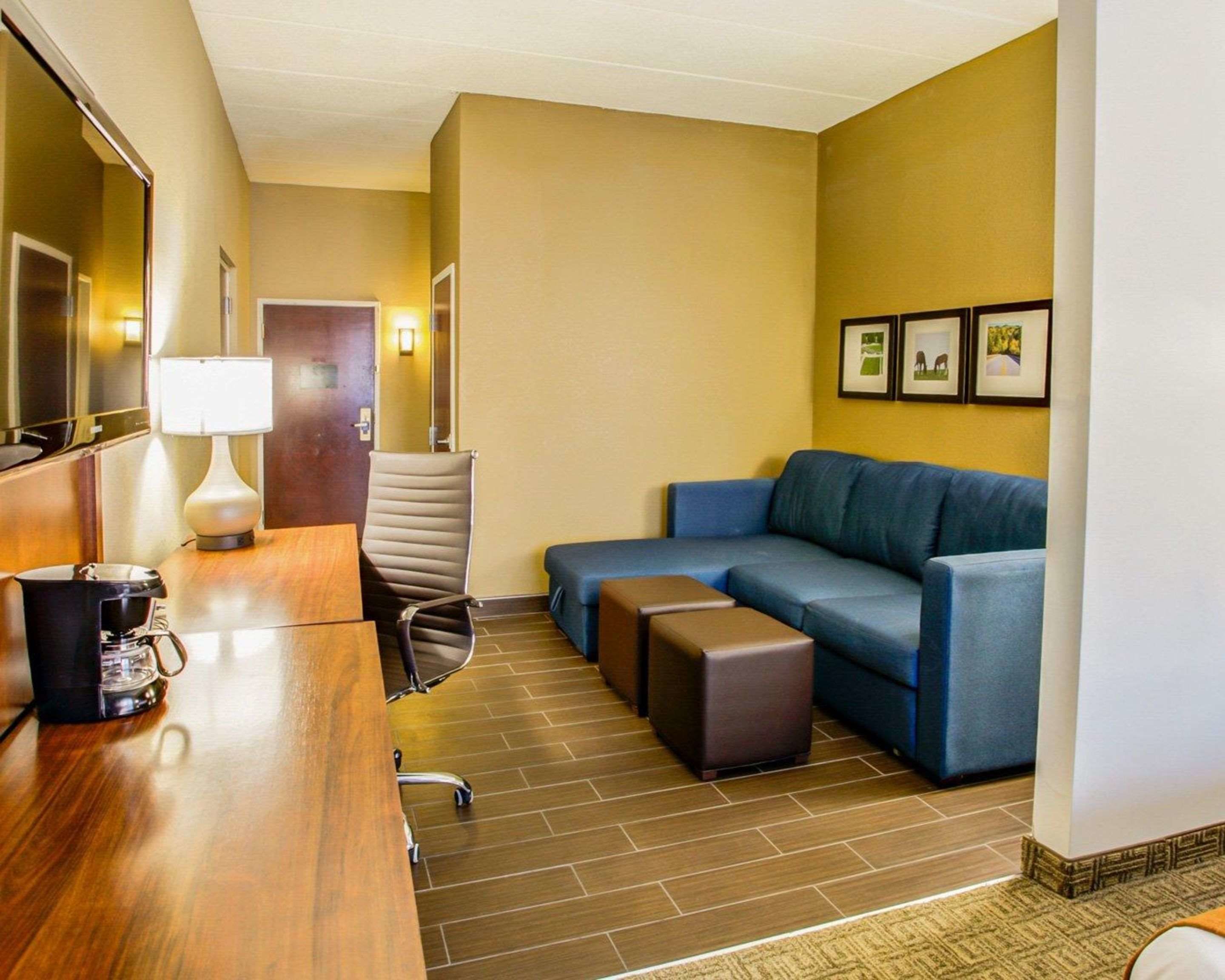 Habitación de hotel con un escritorio de madera, un sofá azul, una silla, cafetera y cuadros en la pared.