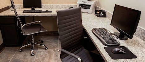 Una oficina de hotel con dos sillas negras, un ordenador de escritorio, una impresora y un monitor.