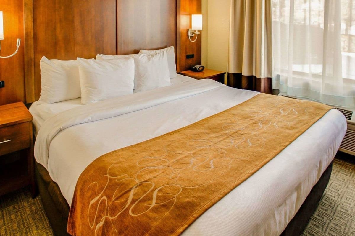 Habitación de hotel con una cama grande, cabecero de madera, dos lámparas de noche, una mesita y una ventana con cortinas.