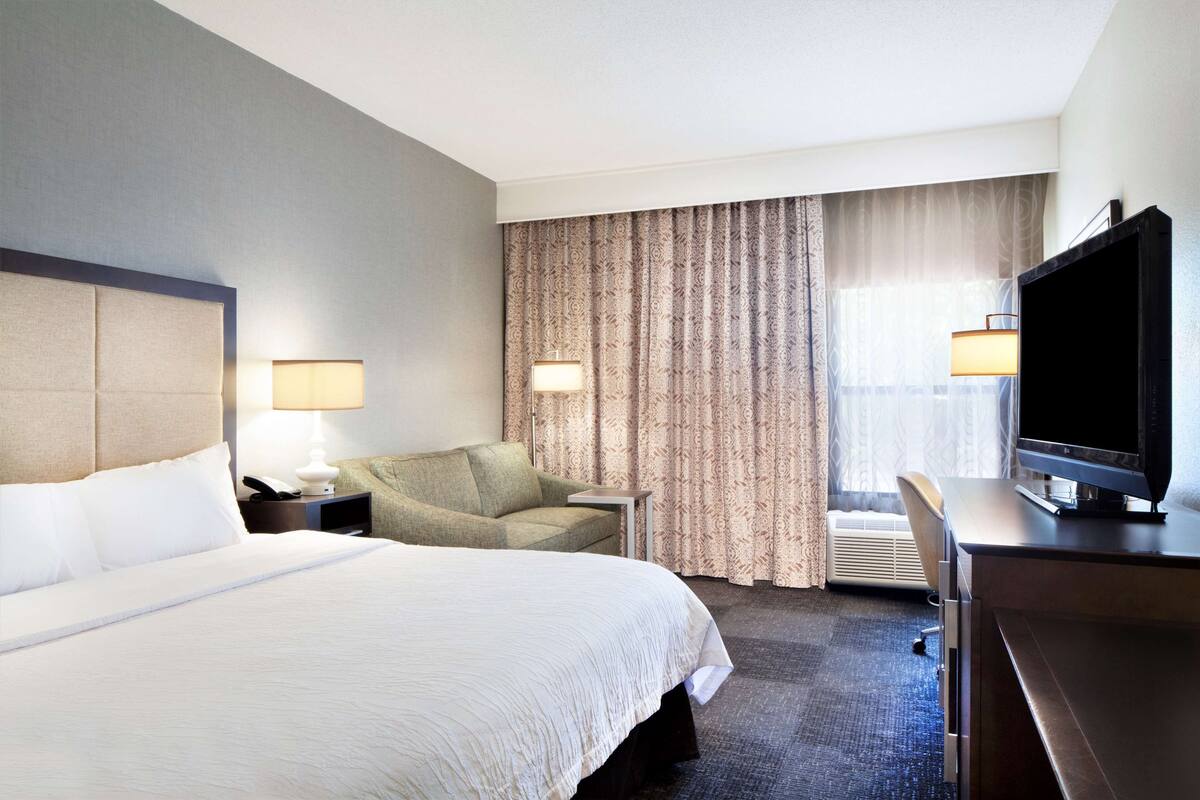 A hotel room with a large bed, a desk with a television, two lamps, a chair, and a window with curtains.