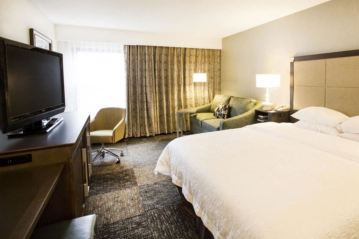 Ein Hotelzimmer mit Bett, Fernseher, Schreibtisch, Stuhl, Nachttisch und Lampe.