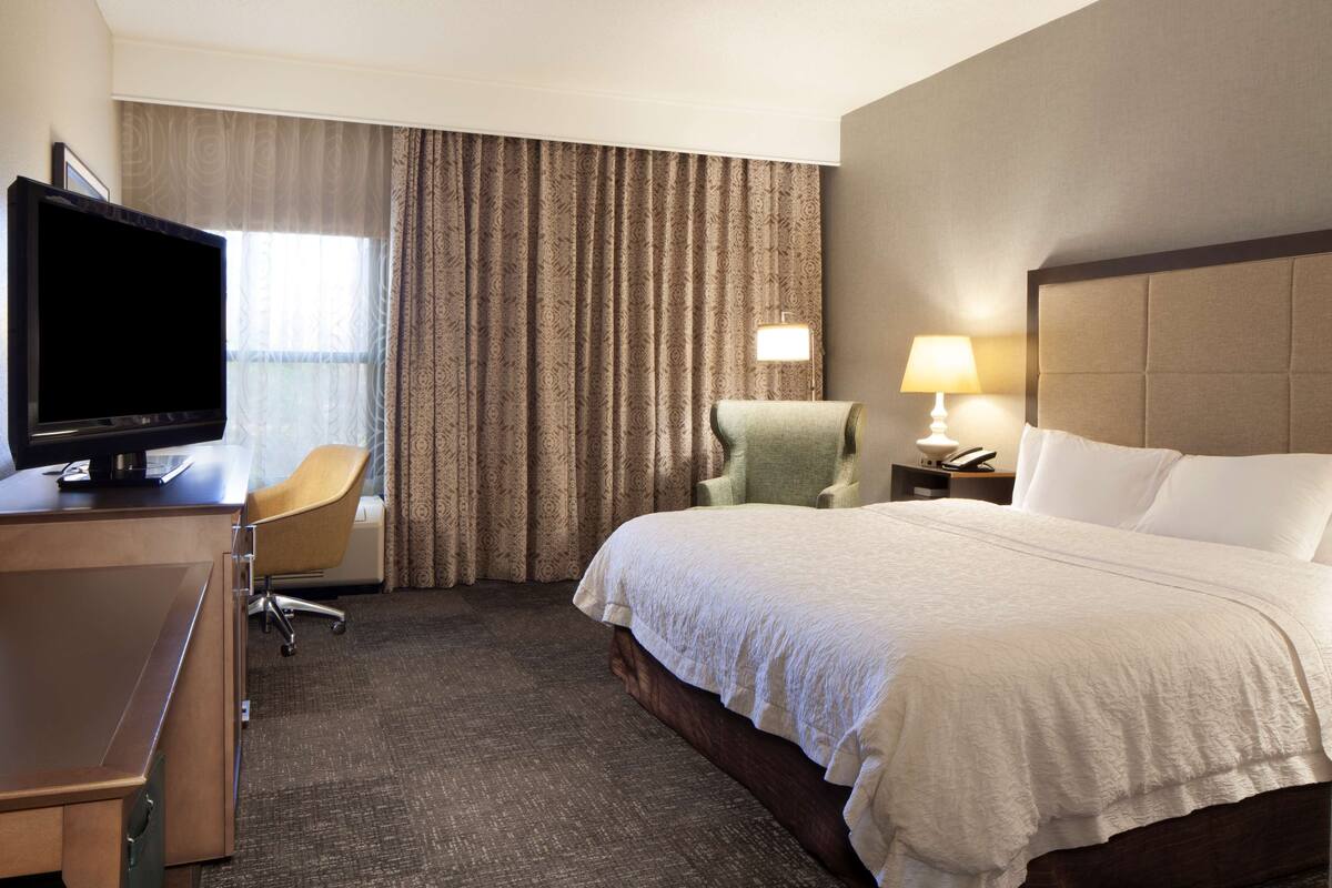 A hotel room with a bed, a desk with a television, a chair, a lamp, and a window with curtains.