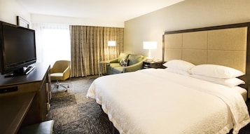 A hotel room with a large bed, a desk with a television, a chair, and a lamp.