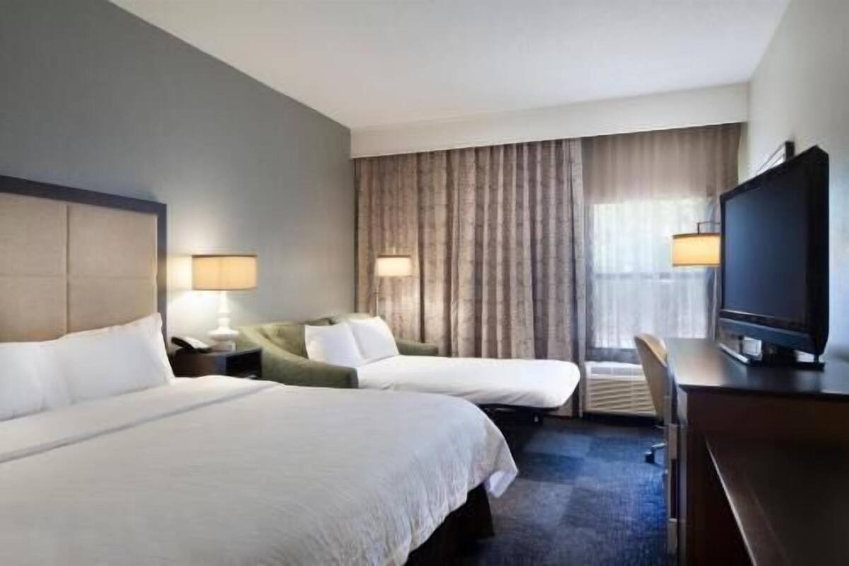 A hotel room with two beds, a desk, a television, and a large window with curtains.