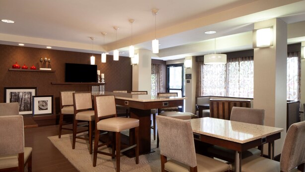 A dining area with wooden tables and chairs, a flat-screen TV mounted on the wall, and a large window with patterned curtains.