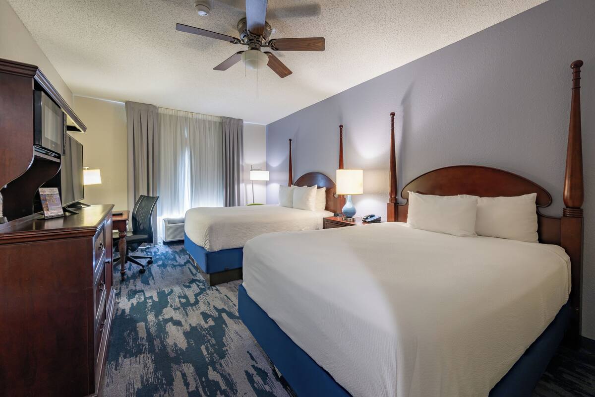 A hotel room with two beds, a desk, a chair, a TV, and a ceiling fan.