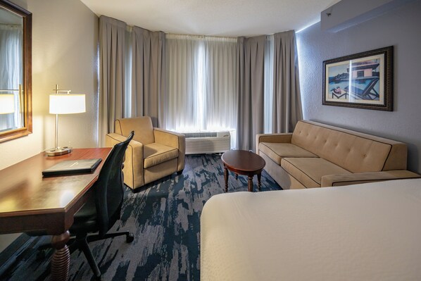 Habitación de hotel con cama, escritorio, silla, sofá, mesita, lámpara, aire acondicionado y ventana con cortinas.