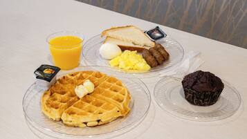 A breakfast tray with waffles, scrambled eggs, sausages, toast, a muffin, and a glass of orange juice.