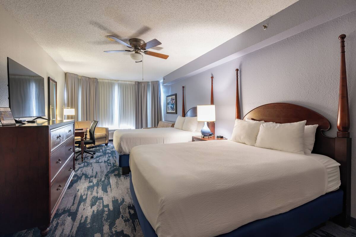 A hotel room with two beds, a ceiling fan, a desk with a chair, and a large window with curtains.