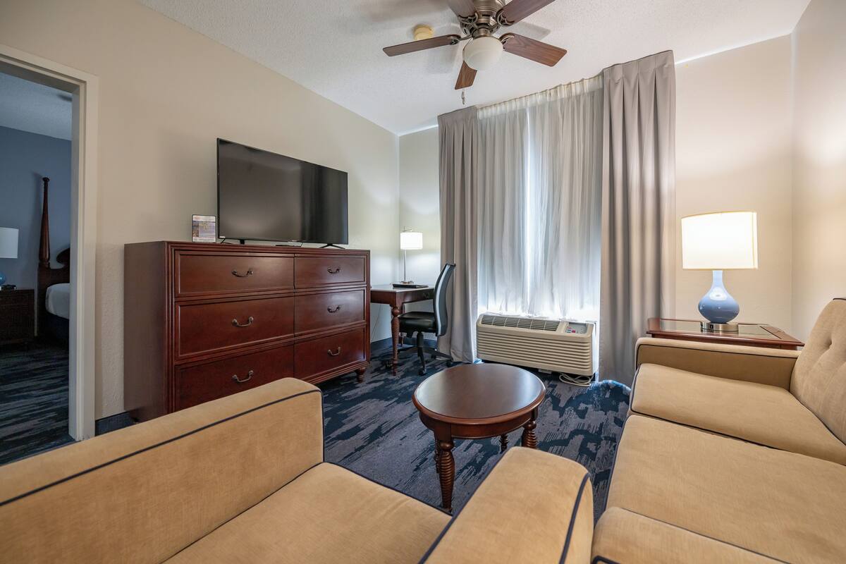 A hotel room with a flat-screen TV, wooden dresser, two sofas, a small table, a ceiling fan, a window with curtains, and a lamp.