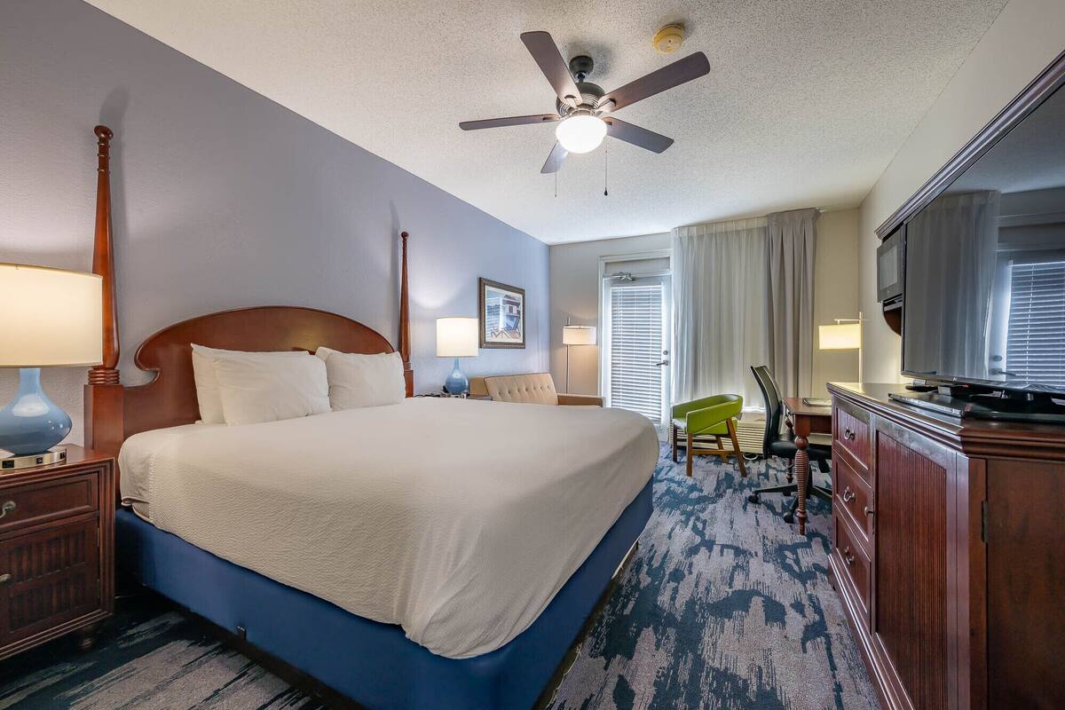 A hotel room with a large bed, a ceiling fan, a TV, and a desk with a chair.