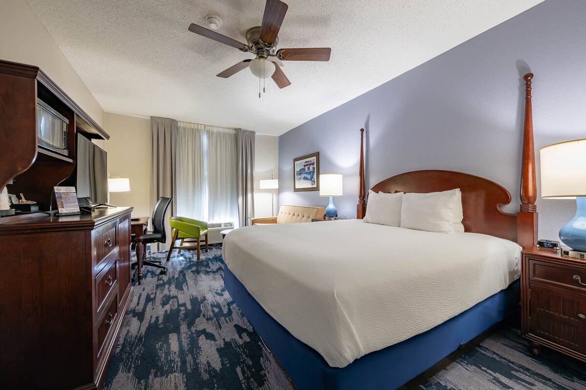 A hotel room with a bed, a desk, a chair, a desk lamp, a nightstand, a clock, and a ceiling fan.