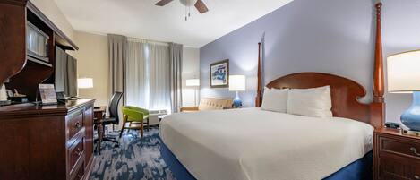 Een hotelkamer met een bed, een bureau, een stoel, een bureaulamp, een nachtkastje, een klok en een plafondventilator.
