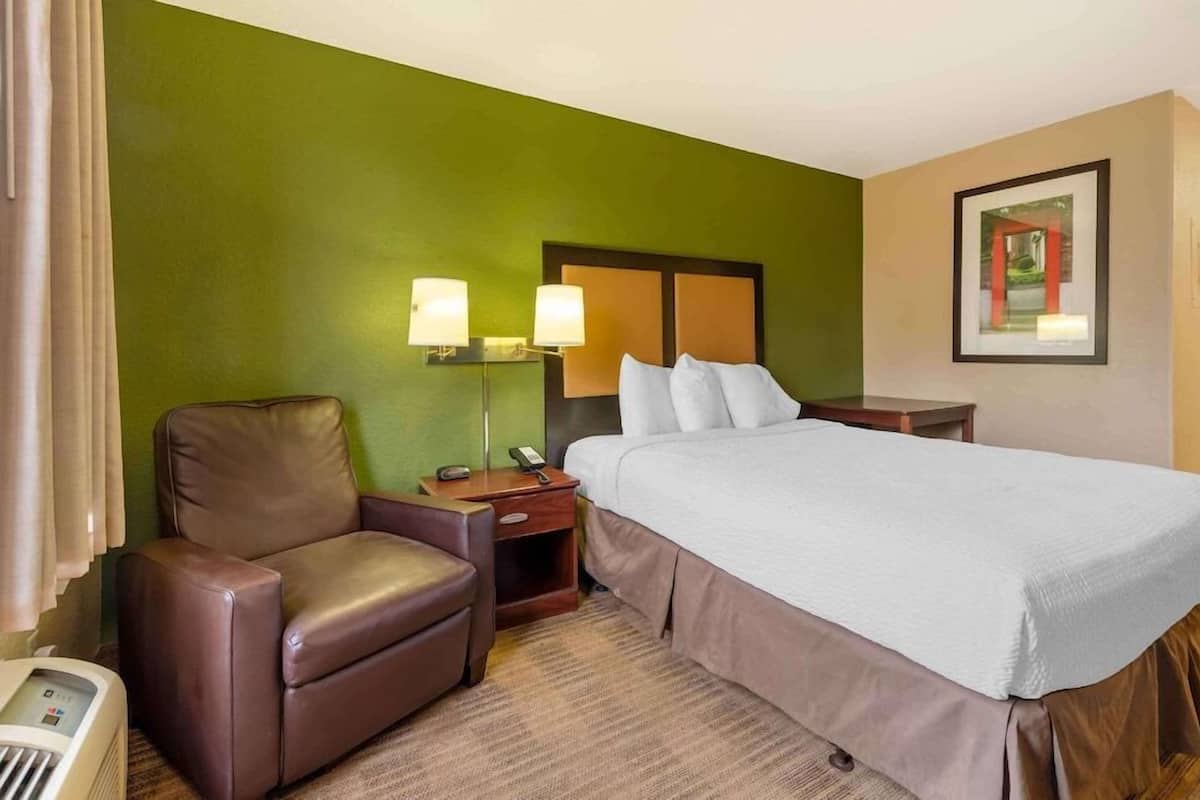 Una habitación de hotel con una cama, una silla, una mesita de noche, una lámpara y un cuadro en la pared.