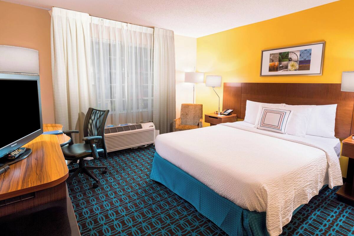 A hotel room with a bed, a desk with a TV, a chair, a lamp, and an air conditioner.