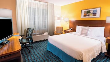 Ein Hotelzimmer mit einem Bett, einem Schreibtisch mit Fernseher, einem Sessel, einer Lampe und einer Klimaanlage.