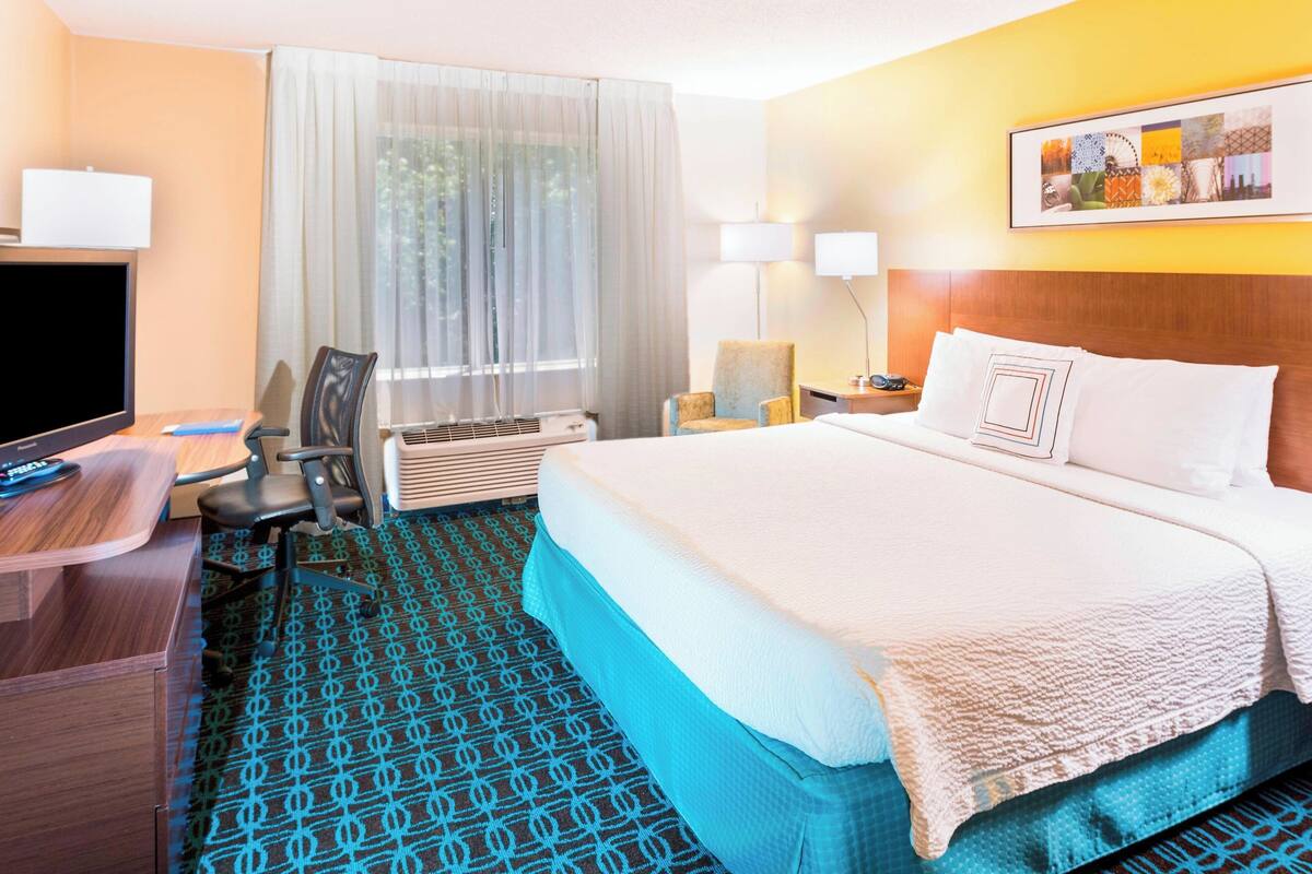 A hotel room with a large bed, a desk with a computer, a chair, a TV, and a window with curtains.