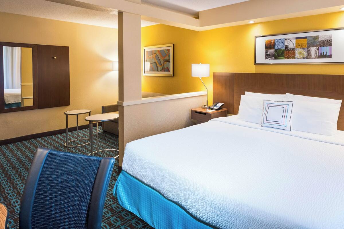 A hotel room with a large bed, a bedside table with a lamp, a small round table, a chair, and a mirror.