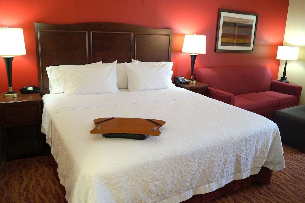 A hotel room with a large bed, two bedside lamps, a red armchair, and a small table with a telephone.