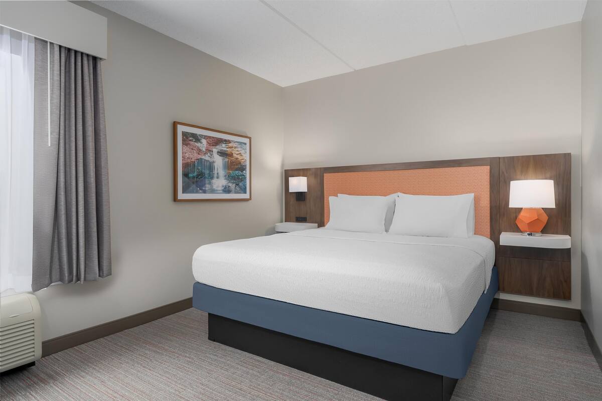 A hotel room with a large bed, a headboard with a light fixture, a bedside table with a lamp, a framed picture on the wall, and a window with curtains.
