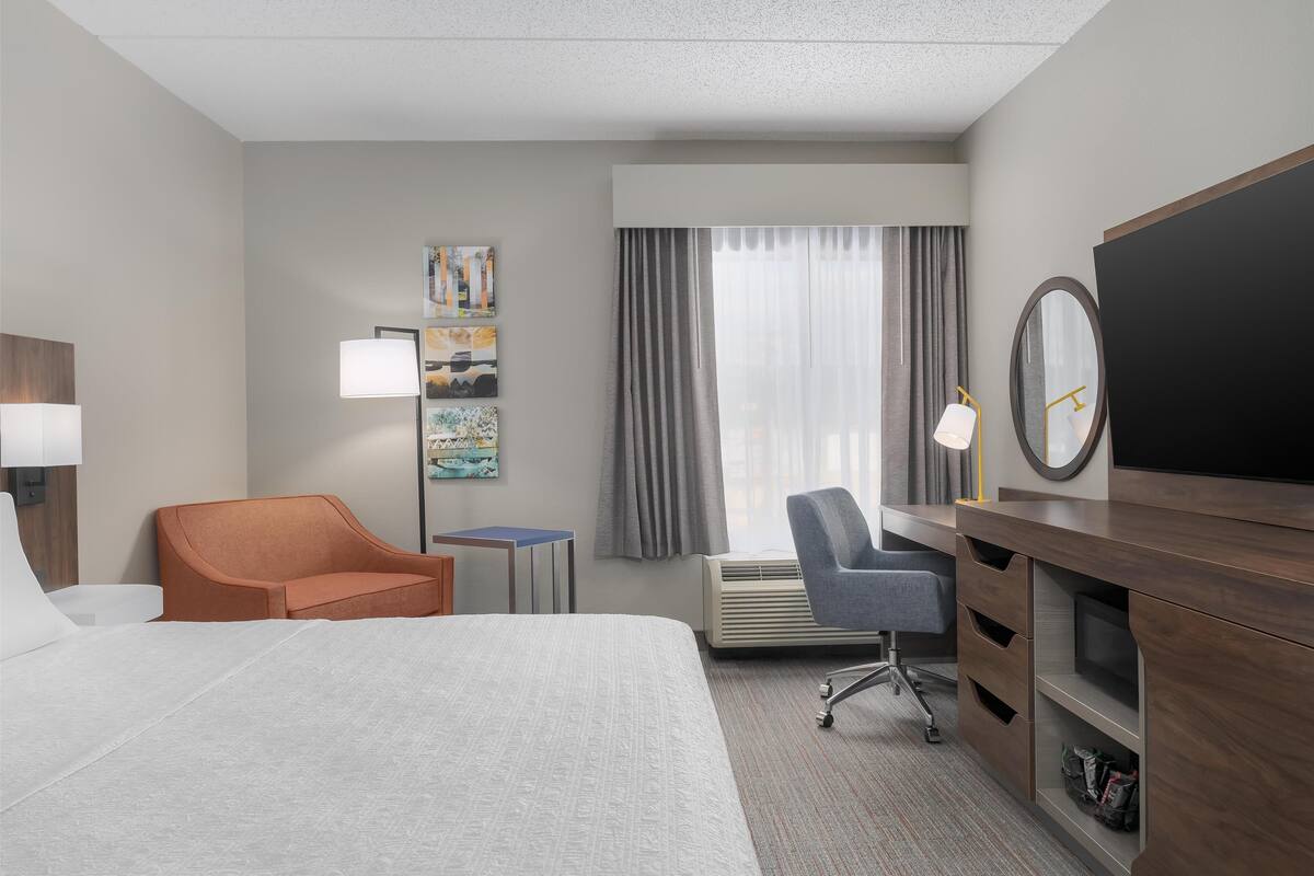 A hotel room with a bed, a desk, a chair, a TV, a mirror, and a window with curtains.