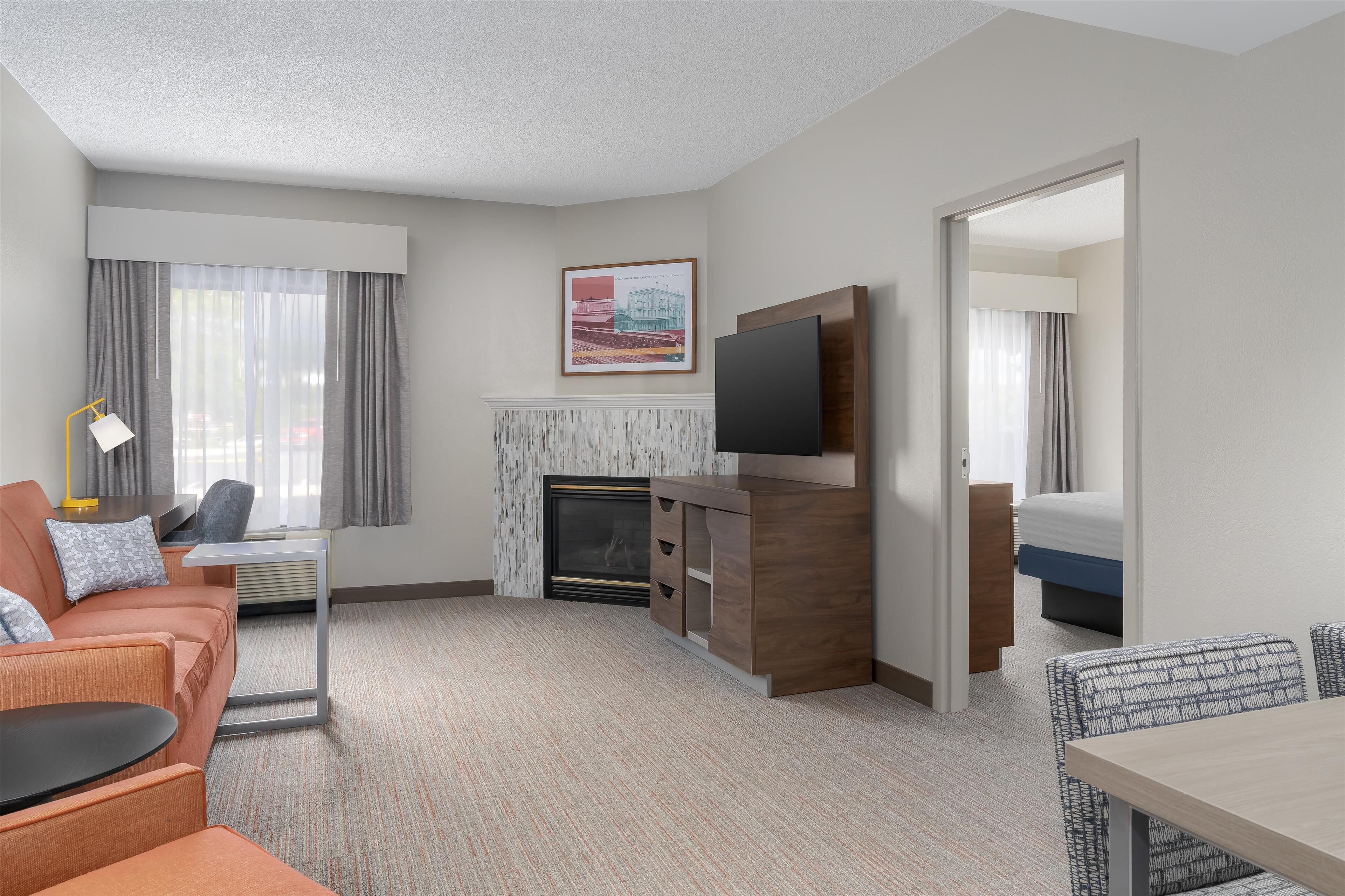 A modern hotel room with a sofa, a small table, a TV mounted on a wooden cabinet, a fireplace, and a bed with a blue headboard.
