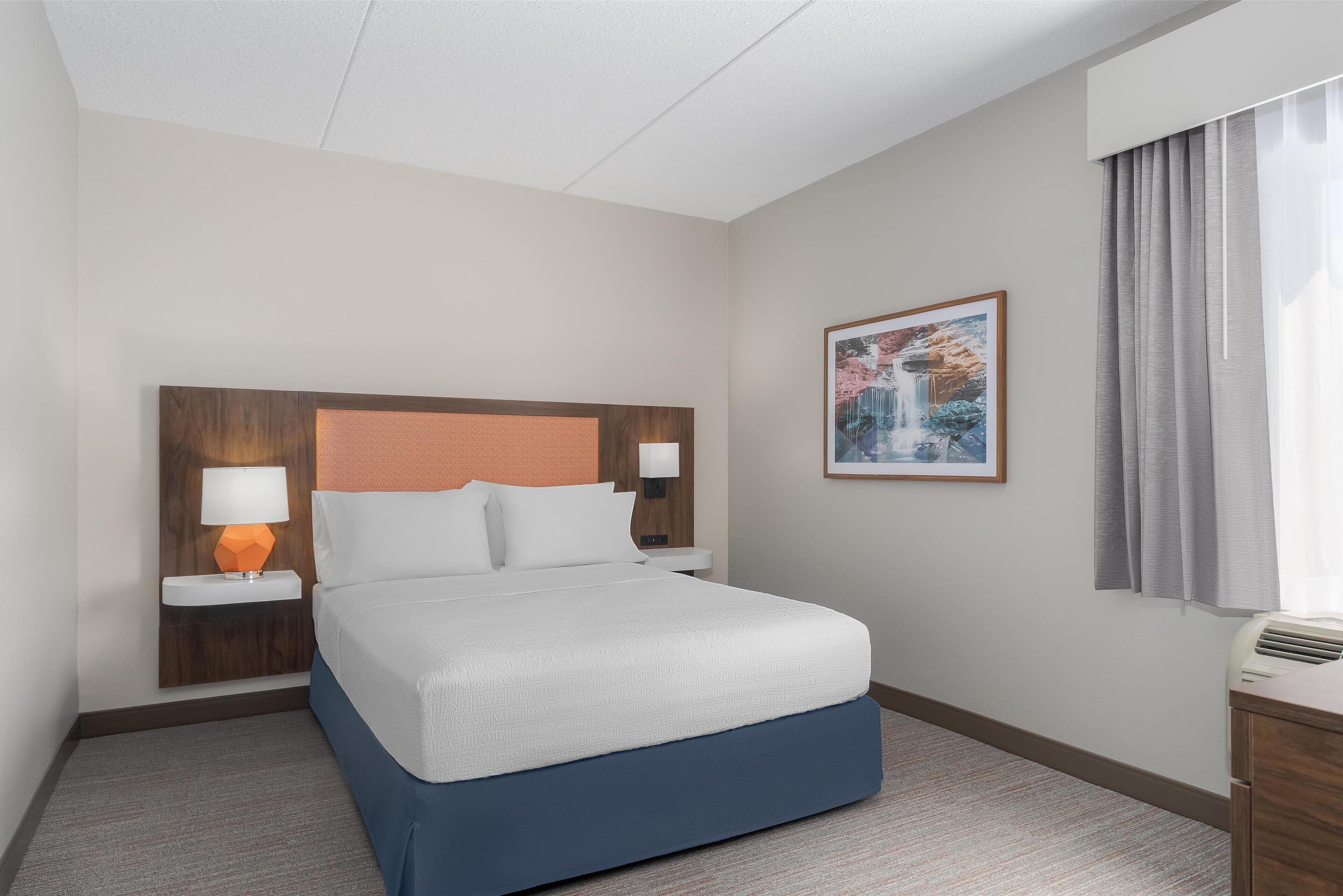 A hotel room with a bed, nightstands, a lamp, a painting, and a window with curtains.