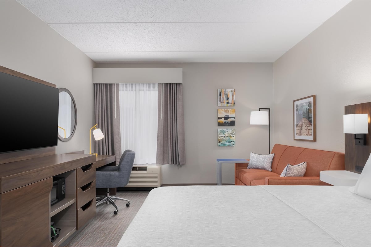 A hotel room with a bed, a desk with a flat-screen TV, a chair, a sofa, and artwork on the walls.