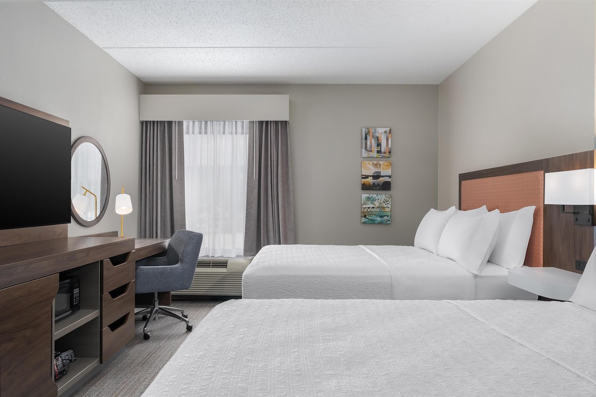 A hotel room with two beds, a desk with a chair, a TV, and a window with curtains.