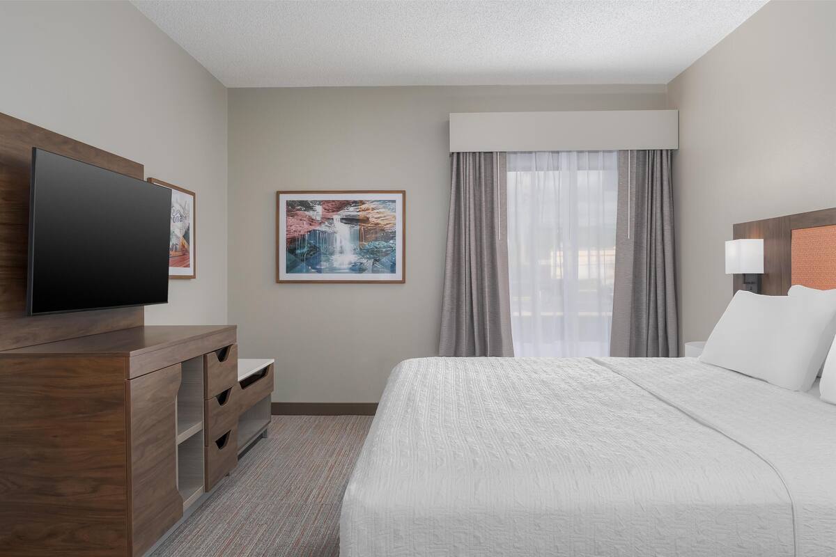 A hotel room with a bed, a TV on a wooden dresser, a painting of a waterfall, and a window with curtains.