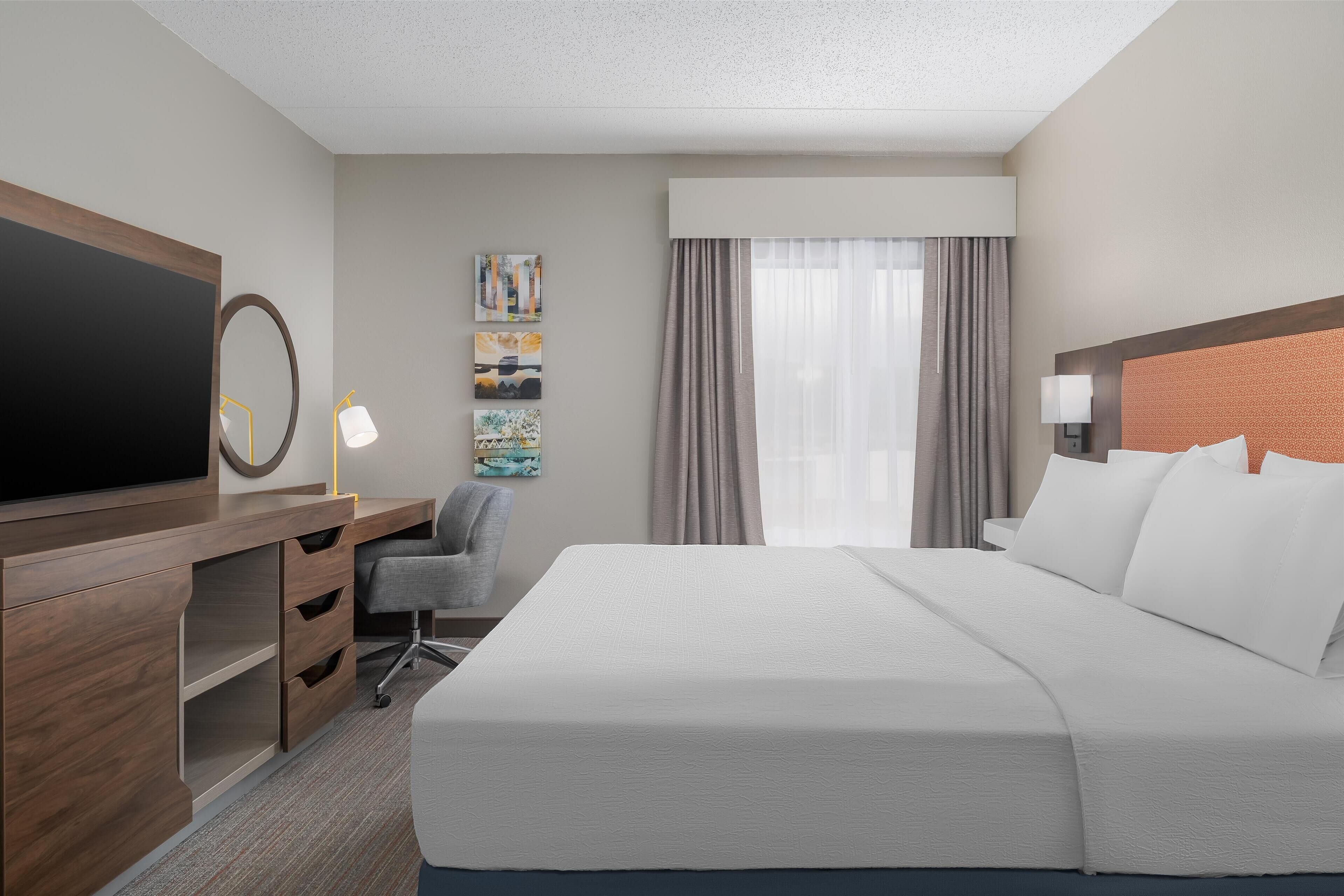A hotel room with a large bed, a desk with a flat-screen TV, a chair, and a window with curtains.