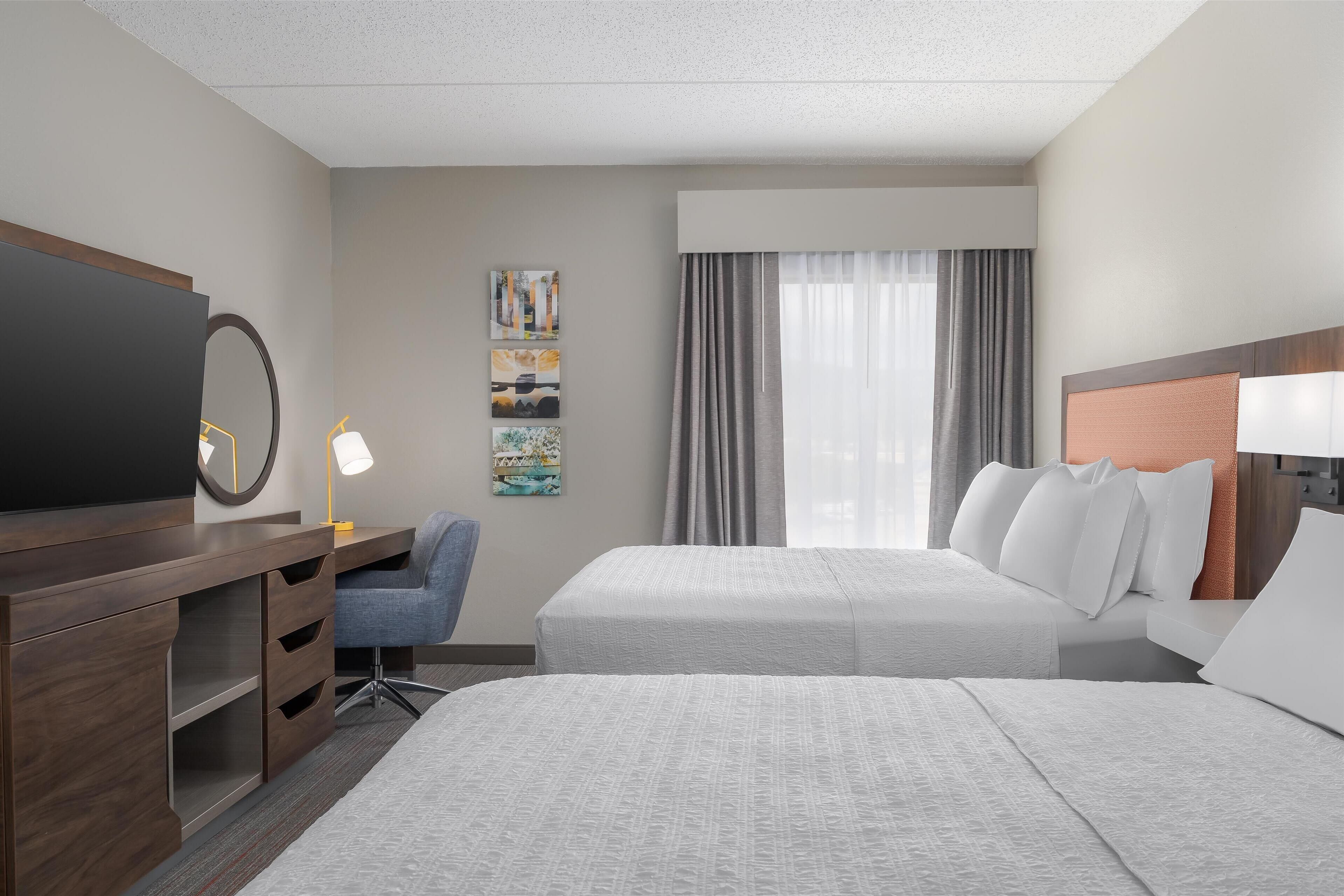 A hotel room with two beds, a desk with a chair, a TV, and artwork on the wall.