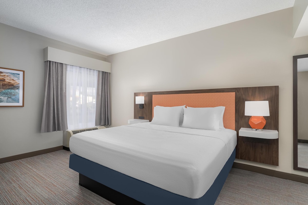 A hotel room with a large bed, a nightstand with a lamp, a window with curtains, and a painting on the wall.