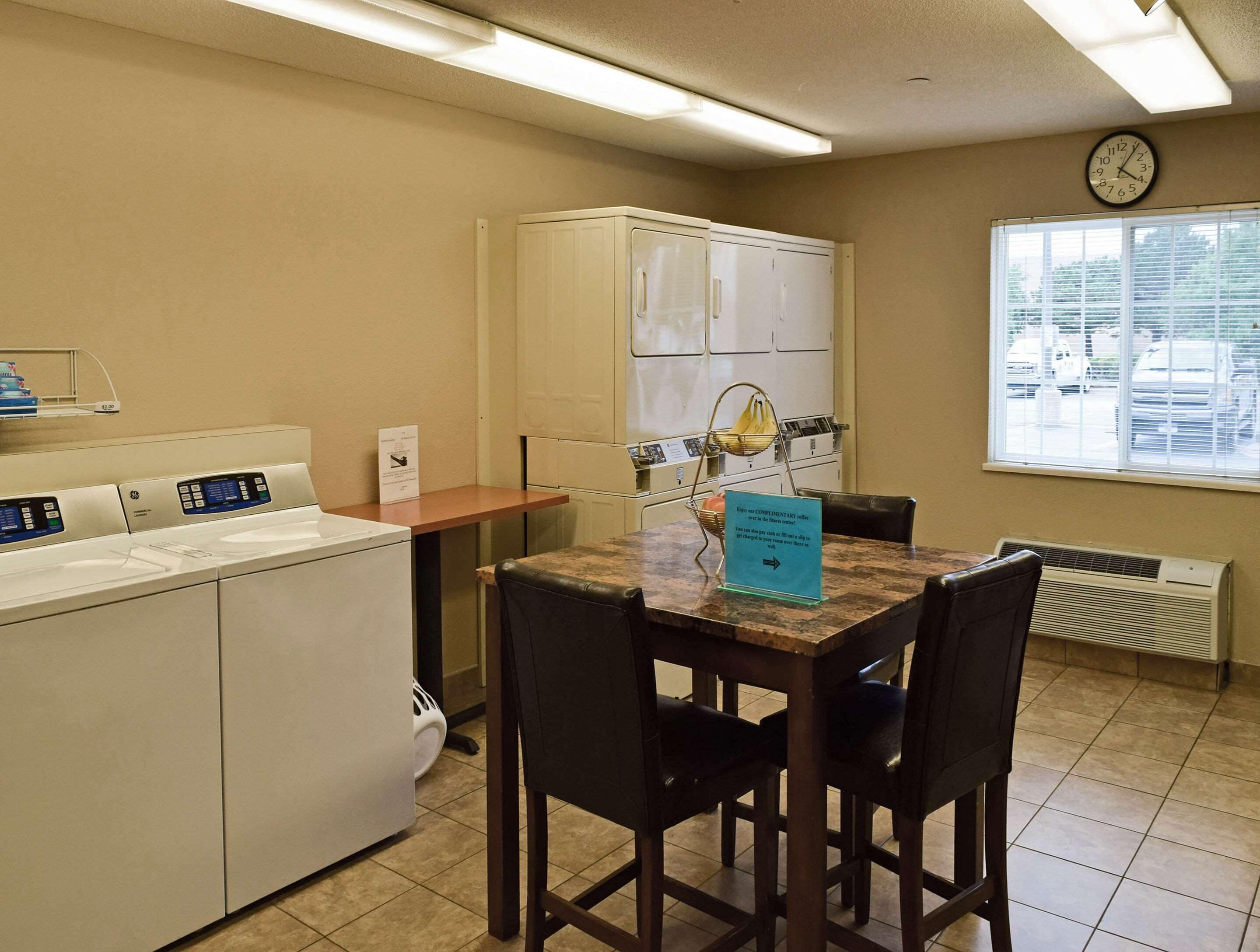 A hotel room with a kitchenette, a dining area with a table and chairs, a window with blinds, and a clock on the wall.