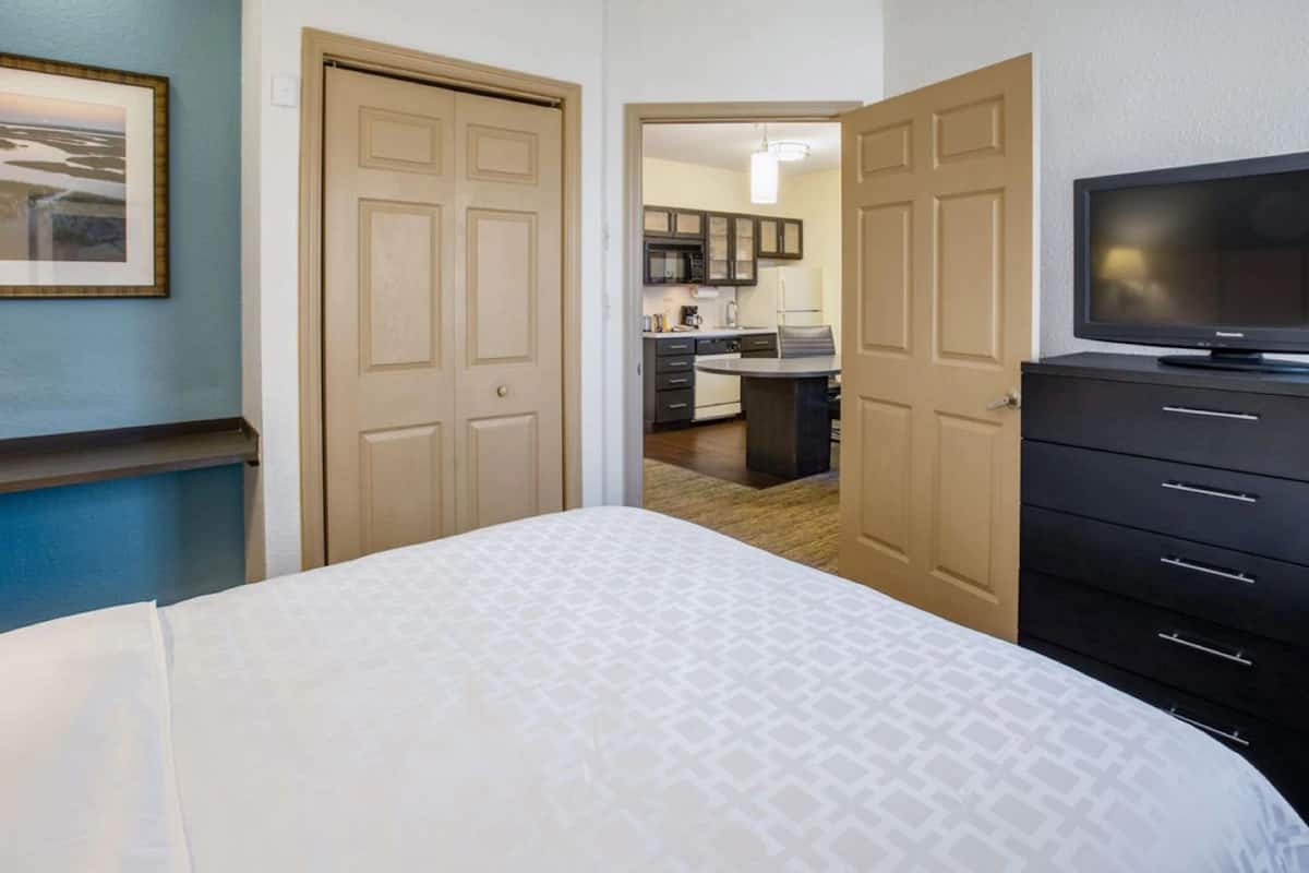 A hotel room with a bed, a TV, a dresser, and a view of the kitchen and dining area.