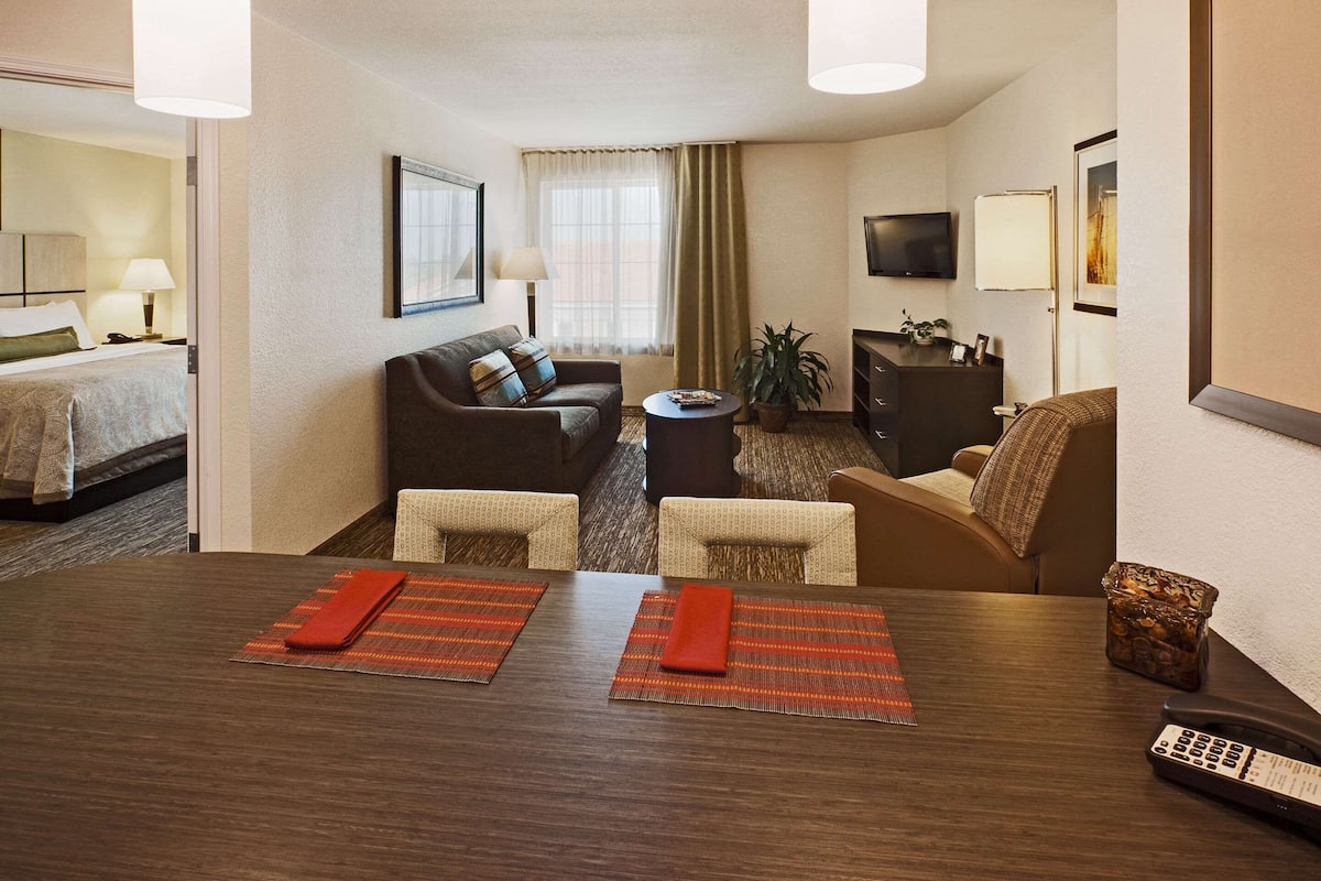 A hotel room with a bed, a sofa, a dining table with placemats, a TV, and a plant.