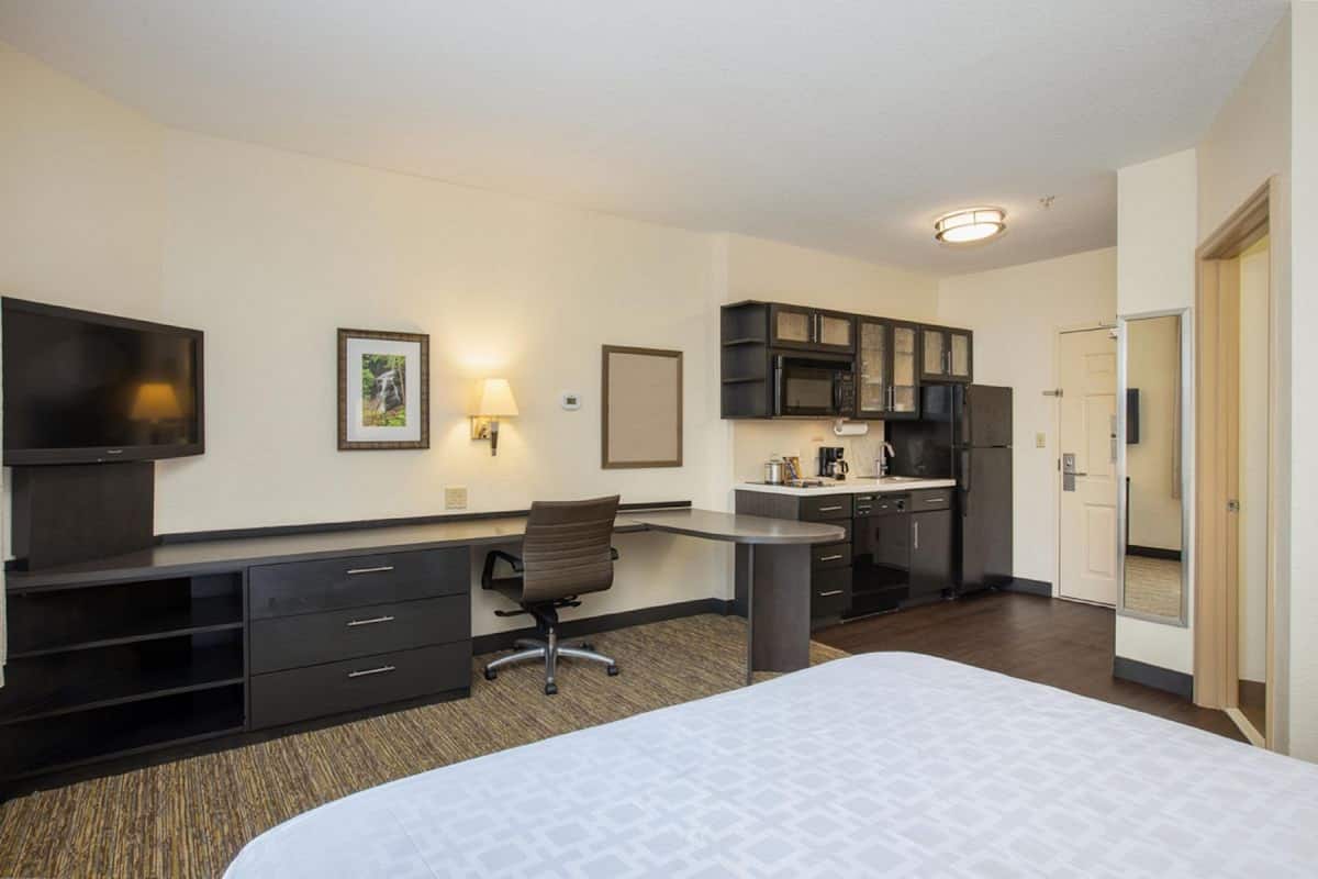 A hotel room with a bed, desk, chair, kitchenette, and a TV.