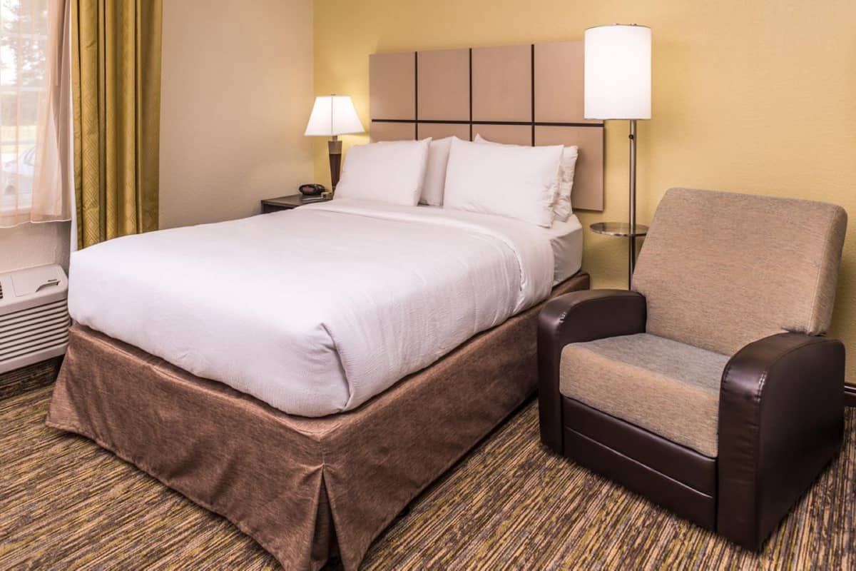 A hotel room with a large bed, a chair, a nightstand, a lamp, and a window with curtains.