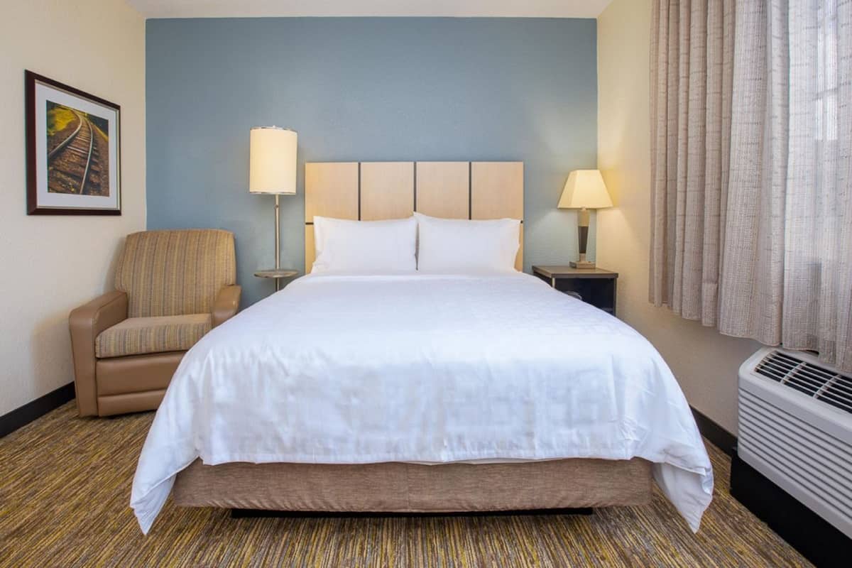 A hotel room with a large bed, two bedside lamps, a chair, a framed picture on the wall, and a window with curtains.