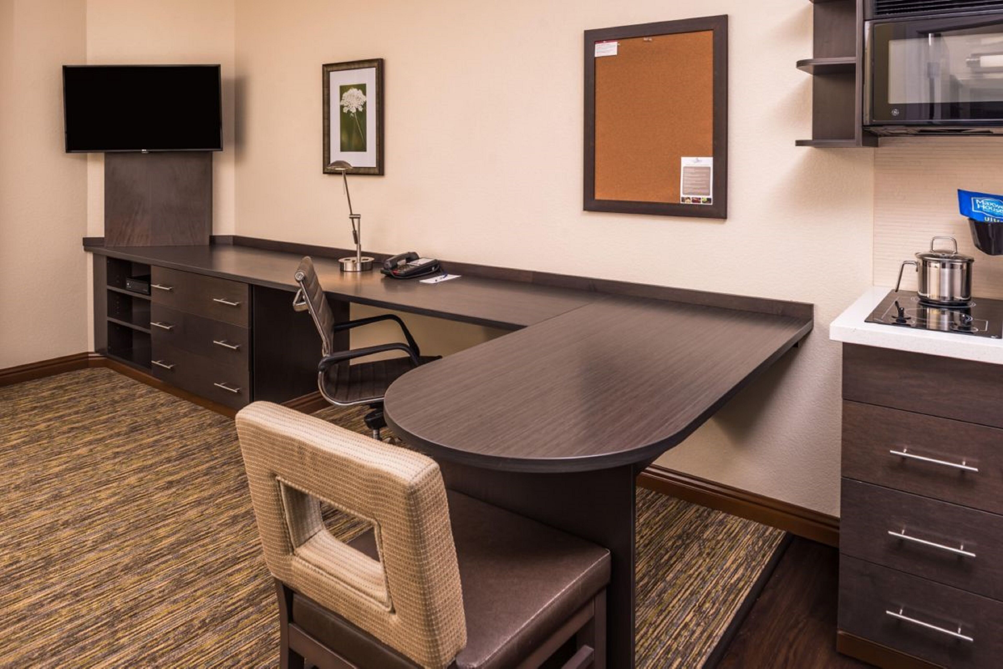 A hotel room with a dark wood desk, a flat-screen TV, a corkboard, a small kitchenette, and a wicker chair.